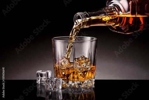 Pouring whiskey from bottle into the cristyl glass with ice cubes on black background.