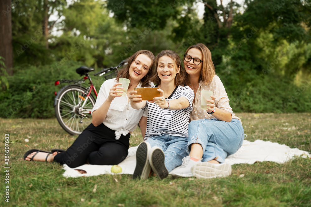 Fototapeta premium Three beautiful happy young women close friends relax in a green park on a blanket. Have fun and chat on the green grass. Taking a selfie on a smartphone