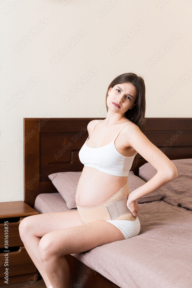 closeup pregnant woman dresses bandage on belly at home on bed. Orthopedic abdominal support belt closeup pregnant woman dresses bandage on belly at home on bed. Orthopedic abdominal support belt