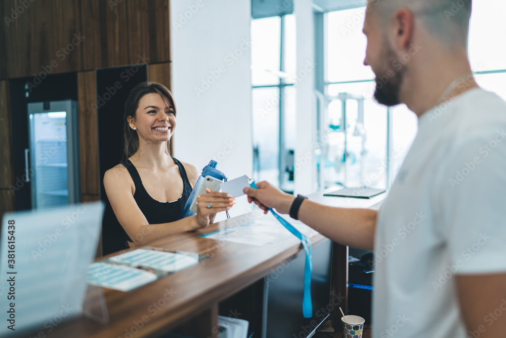 Obraz premium Happy caucasian female in active wear enjoying hospitable service in gym talking with male employee, cheerful 20s woman taking e-key access to locker room having friendly conversation with man