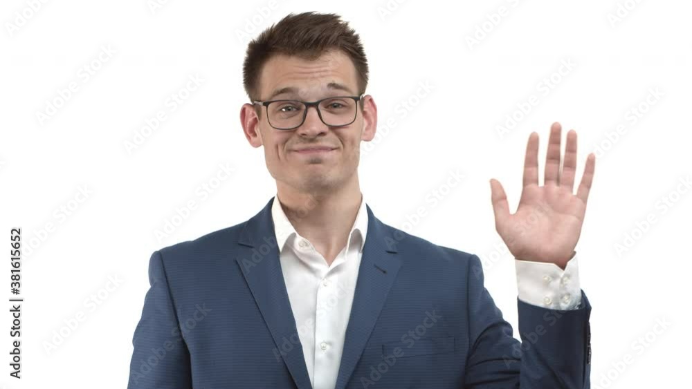 Attractive businessman with blond hair, wearing blue suit and glasses, saying hello and waving hand with friendly smile, standing over white background Attractive businessman with blond hair, wearing blue suit and glasses, saying hello and waving hand with friendly smile, standing over white background