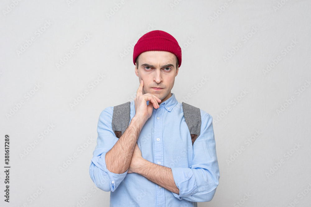 Pensive student with a backpack and a red hat is thinking something, white background, portrait Pensive student with a backpack and a red hat is thinking something, white background, portrait