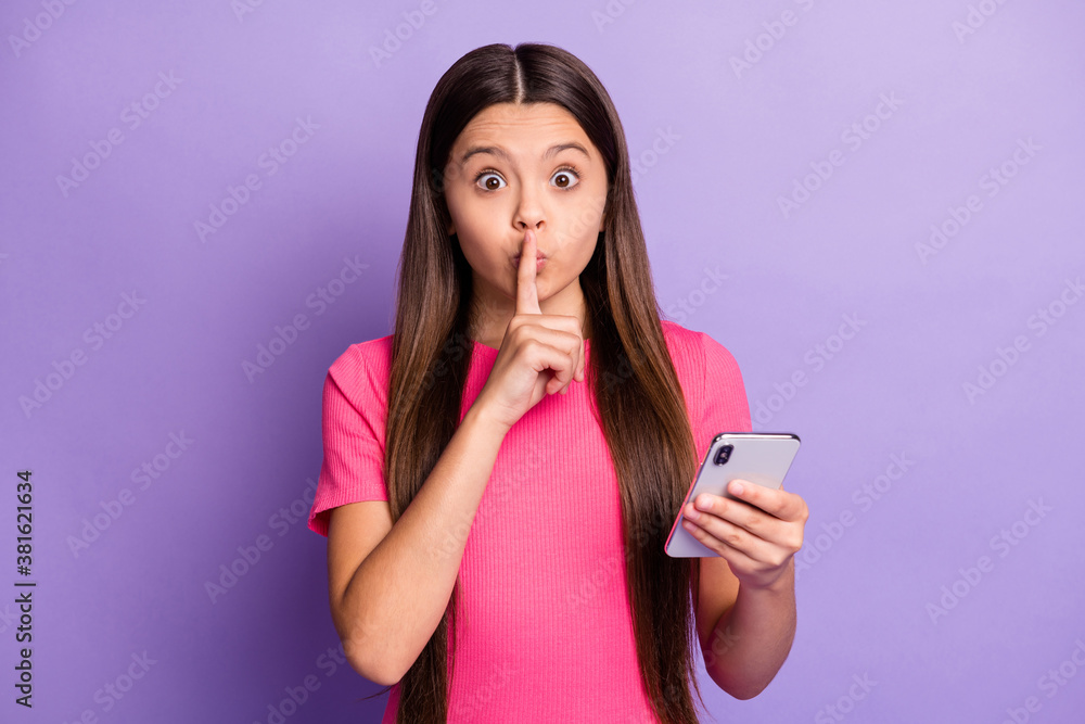Photo portrait of small sweet little girl with long brunette hair keeping secret holding cellphone touching lips with finger isolated on purple color background Photo portrait of small sweet little girl with long brunette hair keeping secret holding cellphone touching lips with finger isolated on purple color background