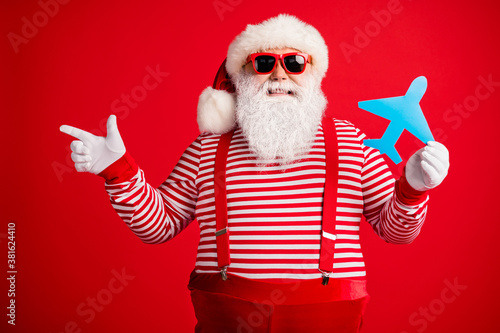 Photo of grandfather grey beard hold blue paper plane direct finger empty space wear santa claus x-mas costume suspenders sunglass striped shirt cap isolated red color background © deagreez