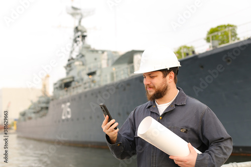 Marine engineer talking by VHF walkie talkie, holding papers near vessel in background.