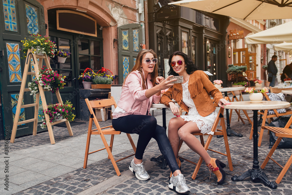 two young beautiful hipster women sitting at cafe, stylish trendy outfit, europe vacation, street style, happy, having fun, smiling, sunglasses, looking at smartphone