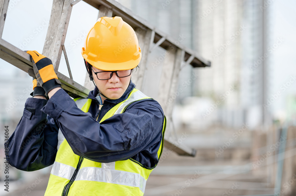 Asian maintenance worker man with safety helmet and reflective suit carrying aluminium step ladder at construction site. Civil engineering, Architecture builder and building service concepts