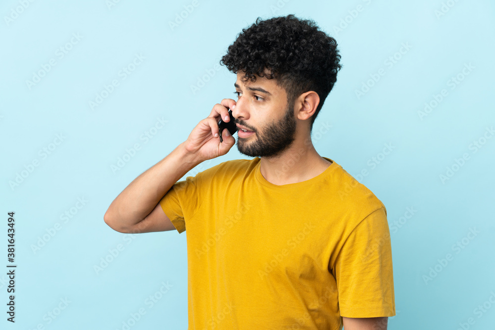 Young Moroccan man isolated on blue background keeping a conversation with the mobile phone with someone Young Moroccan man isolated on blue background keeping a conversation with the mobile phone with someone