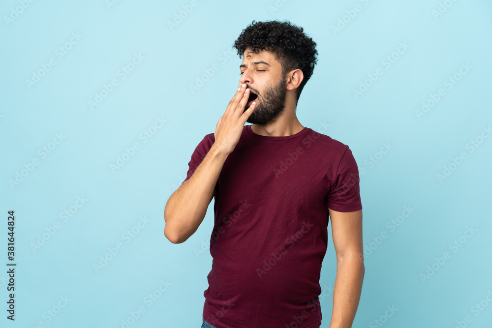 Young Moroccan man isolated on blue background yawning and covering wide open mouth with hand Young Moroccan man isolated on blue background yawning and covering wide open mouth with hand