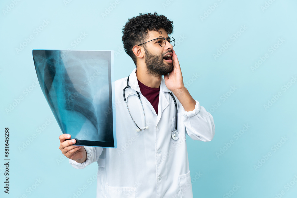 Young Moroccan traumatologist in workplace isolated on blue background shouting with mouth wide open to the side Young Moroccan traumatologist in workplace isolated on blue background shouting with mouth wide open to the side