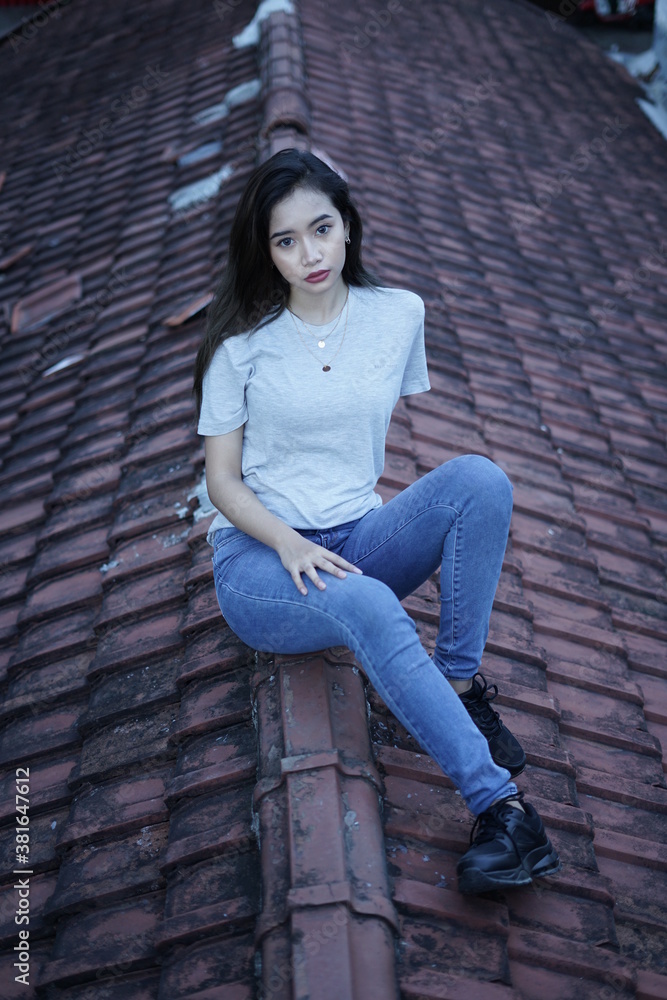 The blonde girl sits on the roof tile while stylishly wearing a gray shirt. female t-shirt models for mockups and templates. The blonde girl sits on the roof tile while stylishly wearing a gray shirt. female t-shirt models for mockups and templates.