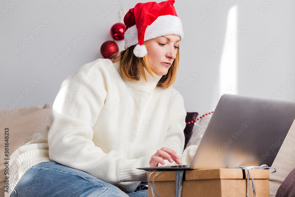 A young woman in a white sweater and Santa Claus hat looks at the laptop screen. Remote work concept A young woman in a white sweater and Santa Claus hat looks at the laptop screen. Remote work concept