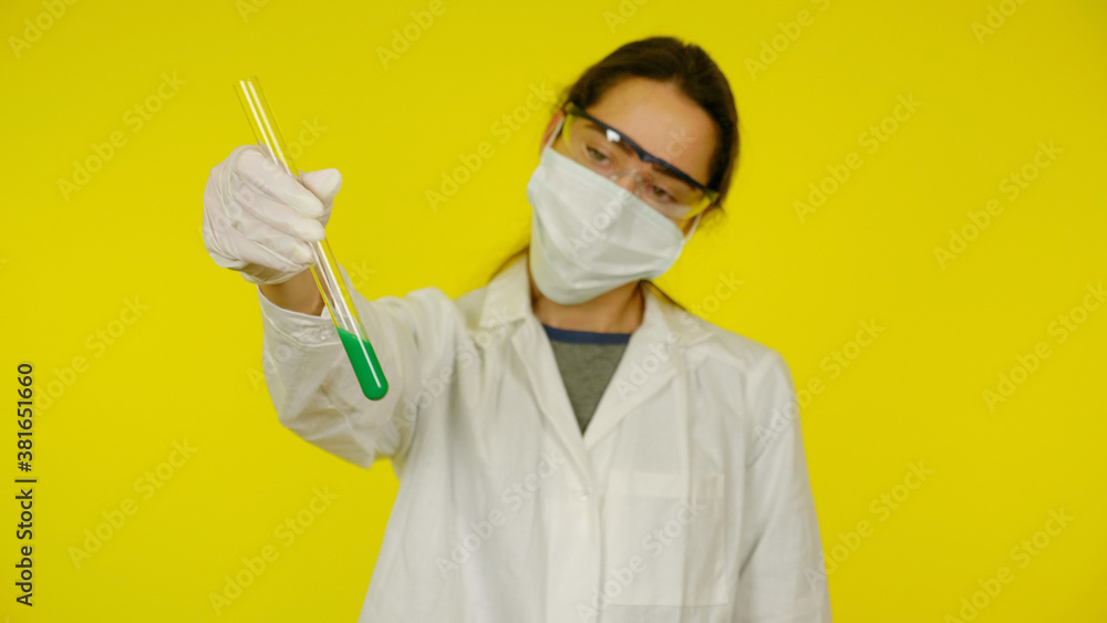 Naklejka premium Young doctor in medical mask goggles latex gloves looks tests in tube. Woman in white coat on yellow background holds hand tube with green liquid