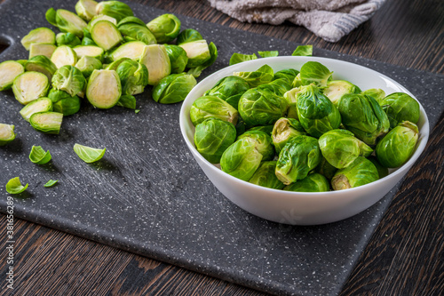 Fresh raw brussels sprouts in a white bowl on a dark cutting board with a towel at the background and wooden table