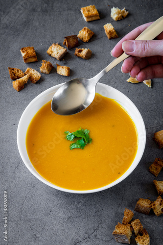 Pumpkin soup with coriander in a white bowl with a spoon coming towards. Croutons and pumpkin seeds in the background. Seasonal autumn / winter food, healthy and fresh