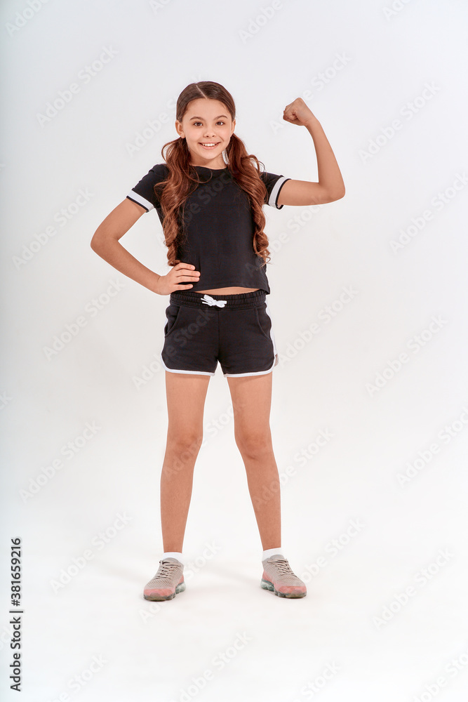 Full length of a cute positive teenage girl in sportswear looking at camera and showing her biceps while posing isolated over grey background in studio Full length of a cute positive teenage girl in sportswear looking at camera and showing her biceps while posing isolated over grey background in studio