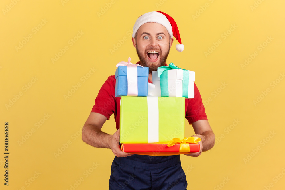 Extremely happy courier in blue uniform and red santa claus hat holding and showing christmas gifts looking at camera with toothy smile. Indoor studio shot isolated on yellow background Extremely happy courier in blue uniform and red santa claus hat holding and showing christmas gifts looking at camera with toothy smile. Indoor studio shot isolated on yellow background