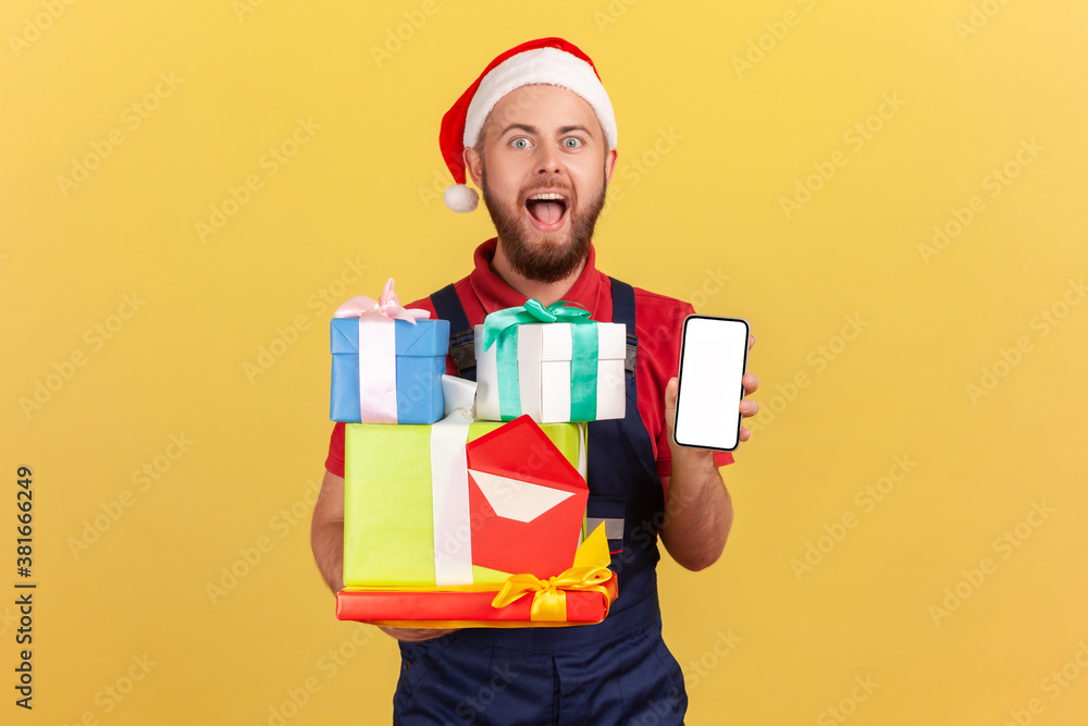 Excited surprised courier in uniform and santa claus hat holding and showing gift boxes and smartphone with empty display, order application. Indoor studio shot isolated on yellow background Excited surprised courier in uniform and santa claus hat holding and showing gift boxes and smartphone with empty display, order application. Indoor studio shot isolated on yellow background