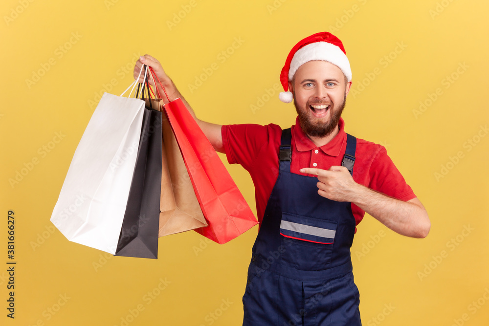 Cheerful positive delivery man in santa claus hat and uniform pointing finger at shopping bags in his hand, fast delivery from store. Indoor studio shot isolated on yellow background Cheerful positive delivery man in santa claus hat and uniform pointing finger at shopping bags in his hand, fast delivery from store. Indoor studio shot isolated on yellow background