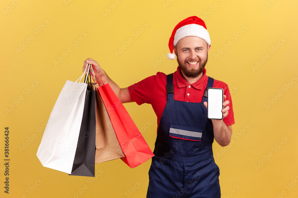 Happy positive courier in santa claus hat and blue uniform holding shopping bags and smartphone, looking at camera with toothy smile, online store. Indoor studio shot isolated on yellow background Happy positive courier in santa claus hat and blue uniform holding shopping bags and smartphone, looking at camera with toothy smile, online store. Indoor studio shot isolated on yellow background