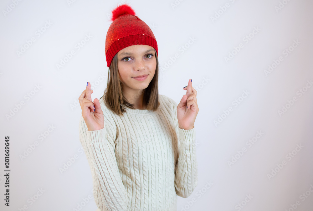 Portrait of a smiling young girl in winter clothes standing over white background, holding fingers crossed for good luck Portrait of a smiling young girl in winter clothes standing over white background, holding fingers crossed for good luck
