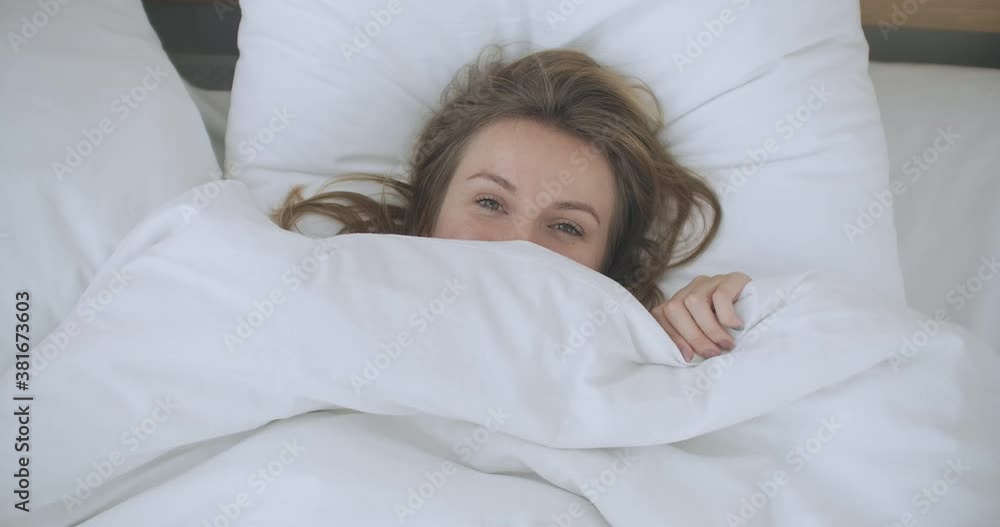 Tanned Smiling Woman Lying In Bed Under A Blanket And Peeks Out From Under Him. Pretty young woman peeking out from under the blanket, smiling at camera. Tanned Smiling Woman Lying In Bed Under A Blanket And Peeks Out From Under Him. Pretty young woman peeking out from under the blanket, smiling at camera.