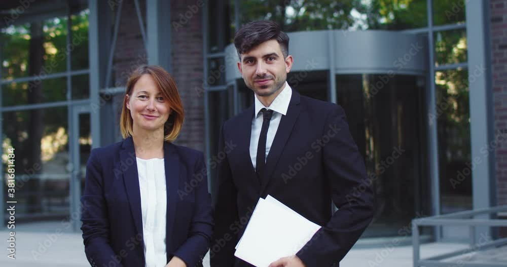 Portrait of confident company workers in black suits outside modern office building. Adult man and woman with documents looking at camera. Business, job, entreprenuership concept. Portrait of confident company workers in black suits outside modern office building. Adult man and woman with documents looking at camera. Business, job, entreprenuership concept.