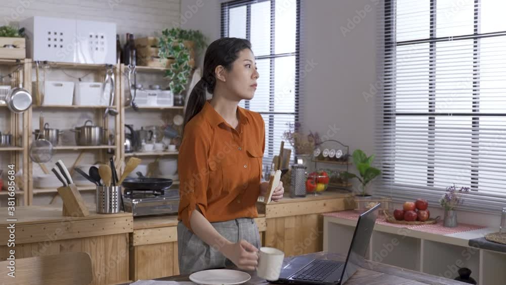 asian woman dressed in business casual is standing while having bread and sipping coffee in the morning at home, looking into distance and thinking about something. asian woman dressed in business casual is standing while having bread and sipping coffee in the morning at home, looking into distance and thinking about something.