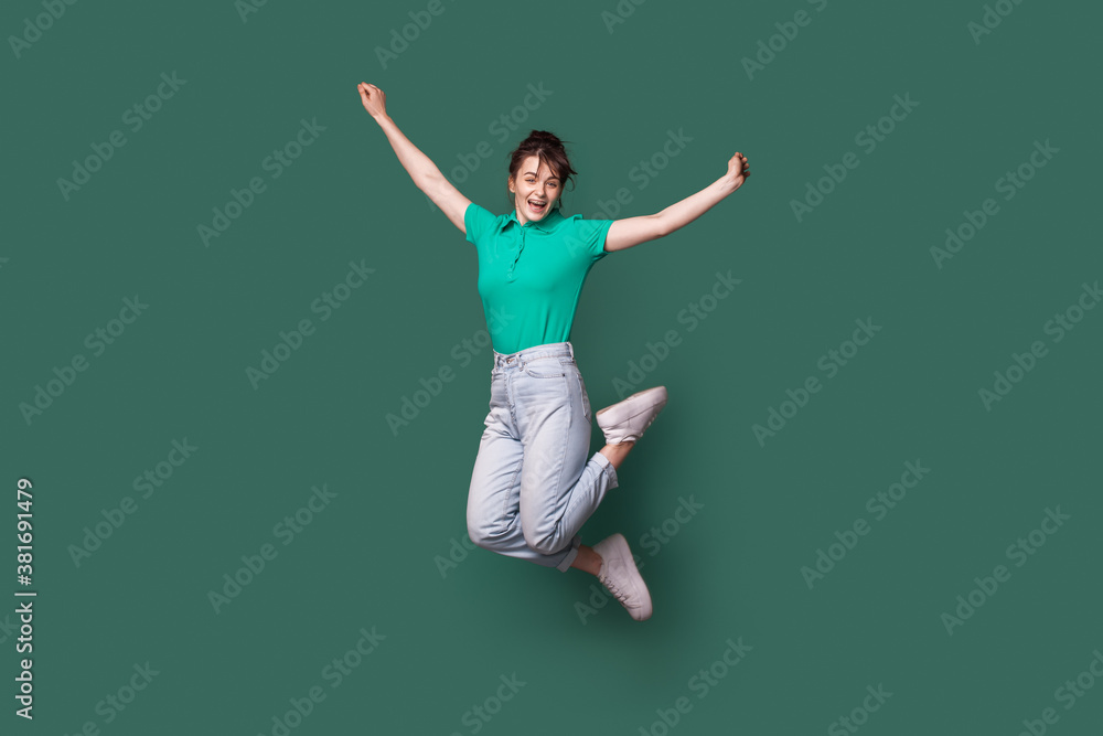 Brunette caucasian woman in jeans and t-shirt jumping on a green studio wall and smile