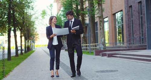 Adult man and woman walking outside modern office building and discussing project. Pretty business lady showing charts to male collegue with laptop. Partnership, corporation, company. photo
