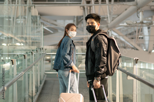 Two Asian traveller couple in casual clothes wearing face mask to prevent coronavirus walking with luggage in airport walkway. new normal and keep social distancing lifestyle. safety travel concept