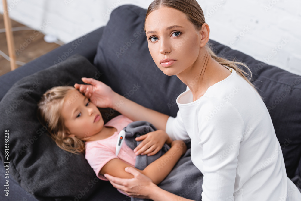 Selective focus of woman looking at camera while touching forehead of daughter with thermometer Selective focus of woman looking at camera while touching forehead of daughter with thermometer