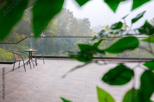 Autumn fog in the old majestic park. Colorful leaves on the trees. Glass panels on the edge of the terrace. Two garden chairs and a table on the summer terrace.