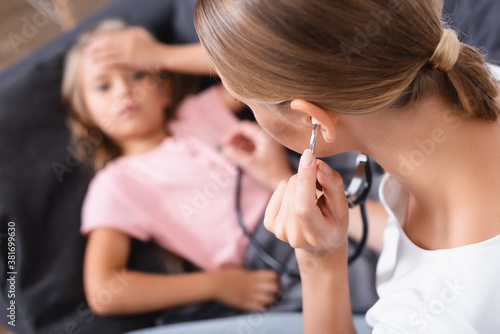 Selective focus of mother in stethoscope sitting beside ill daughter on couch
