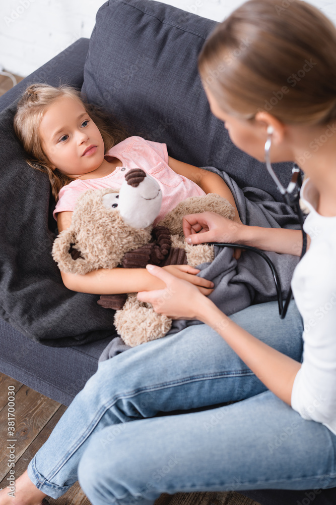 Selective focus of woman in stethoscope sitting near daughter with teddy bear on sofa Selective focus of woman in stethoscope sitting near daughter with teddy bear on sofa