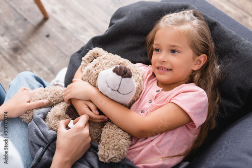 Selective focus of woman holding stethoscope near daughter embracing teddy bear at home