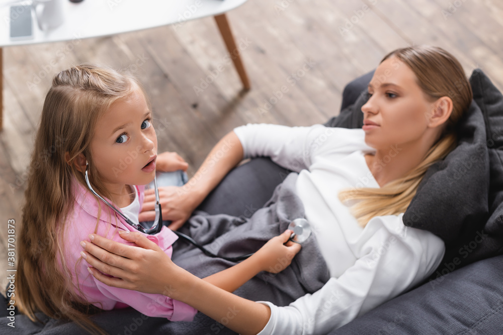 High angle view of child in stethoscope looking at camera near sick mother in living room High angle view of child in stethoscope looking at camera near sick mother in living room