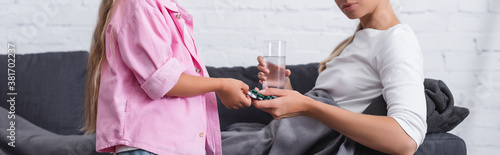 Panoramic shot of child giving pills to sick woman with glass of water on sofa