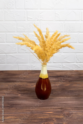 A bouquet of dry spikelets in a brown ceramic vase on a wooden table against a white brick background. Stylish home decor. Modern interior design photo