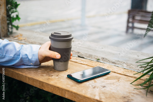 Photo of a reusable cup of coffee on a wood table with the phone. A hand with casual clothes is holding the cup. Job break. Smart working