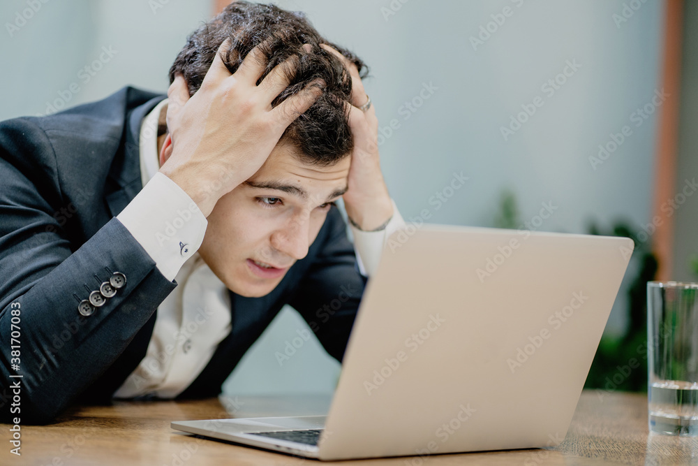 holding his head strongly worried male businessman of European appearance in a stylish black suit looking at a laptop monitor. holding his head strongly worried male businessman of European appearance in a stylish black suit looking at a laptop monitor.