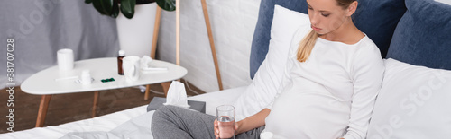 Panoramic shot of pregnant woman sitting on bed while holding glass of water and jar of pills