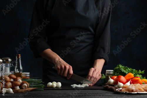 The chef in black uniform cuts with knife onion on black board for adds to the meal. Dark blue background. Preparing food concept.