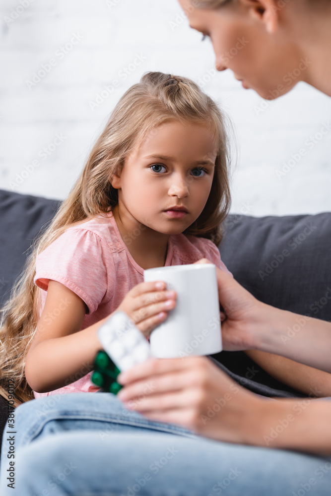 Selective focus of child looking at camera near mother with mug and pills at home
