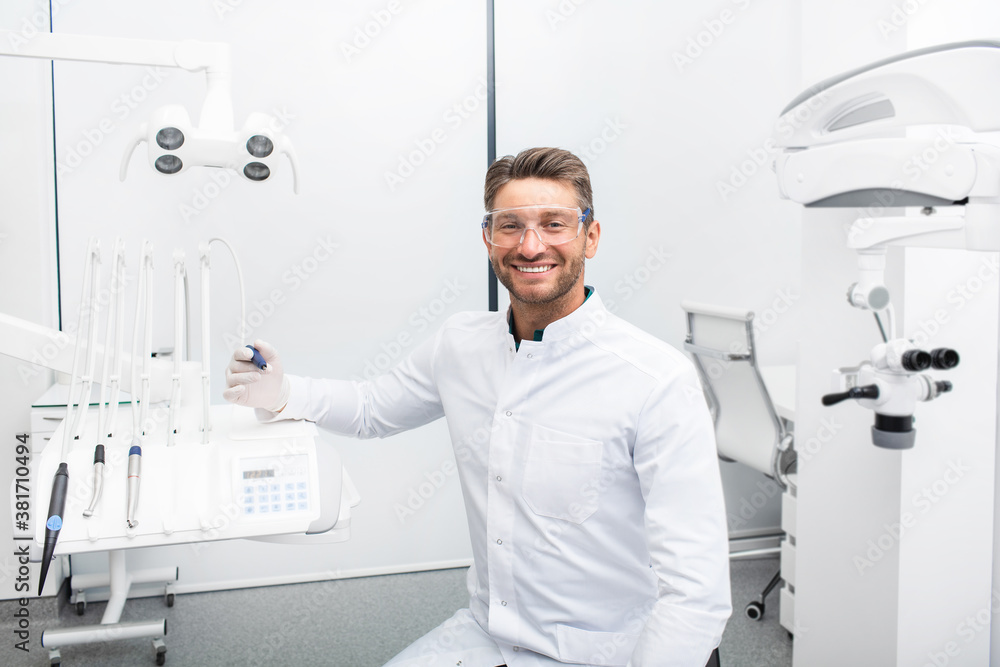 Confident male dentist sitting in a modern clinic near his dental chair. Dental hygienist wearing protective glasses smiling at the camera Confident male dentist sitting in a modern clinic near his dental chair. Dental hygienist wearing protective glasses smiling at the camera