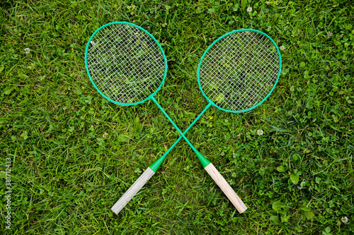 Close up view of badminton rackets laying on green grass crossed as x, outdoors nature background. Badminton game, sport, activity, summer, hobby, competition, tournament concept. Copy text space