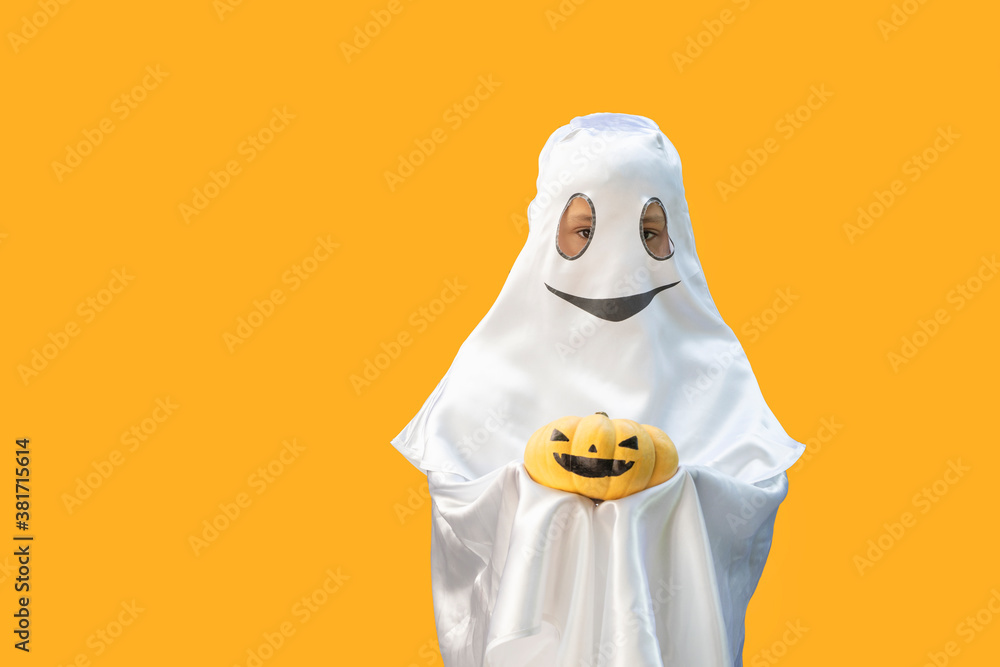 little boy looking out from ghost costume with smile holding orange pumpkin  with black painted face on  orange background. Halloween celebration concept. Copy space for text.