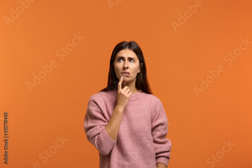 Difficult decision. Image of hesitant beautiful woman in pink sweater, holds finger on chin, bites lip, doubts what to choose over orange background