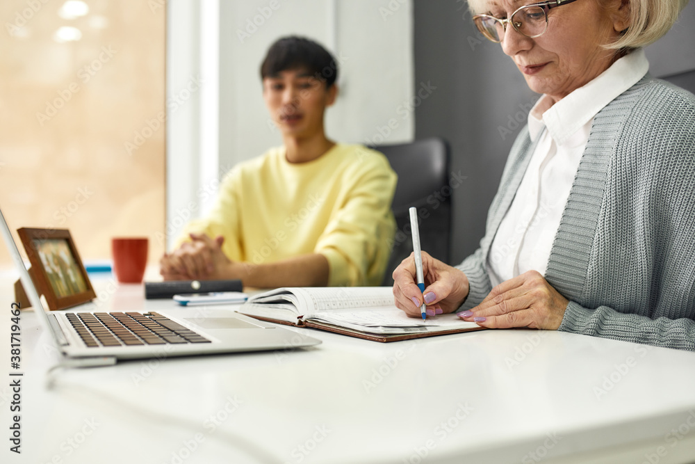Cropped shot of aged woman, senior intern making notes while listening to her young colleague, Friendly male worker training or teaching new employee Cropped shot of aged woman, senior intern making notes while listening to her young colleague, Friendly male worker training or teaching new employee