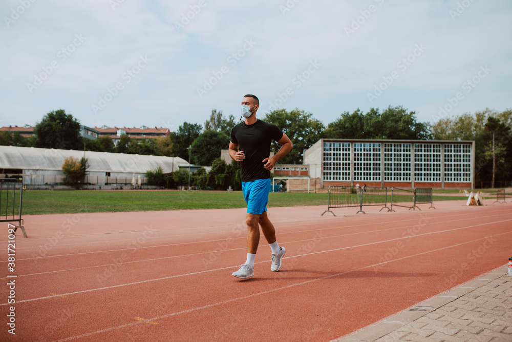 A shot of a young male caucasian athlete  with face mask training on a racetrack. The sprinter runs on athletic all-weather running track. COVID - 19 coronavirus protection